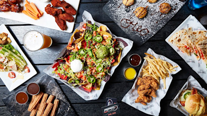 A top view of various dishes on a dark table, including a plate of nachos topped with jalapeños, guacamole, and sour cream, a quesadilla, seasoned french fries, chicken wings, and a glass of beer. Smaller plates hold desserts, fried items, and dipping sauces.