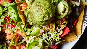 A top view of various dishes on a dark table, including a plate of nachos topped with jalapeños, guacamole, and sour cream, a quesadilla, seasoned french fries, chicken wings, and a glass of beer. Smaller plates hold desserts, fried items, and dipping sauces.