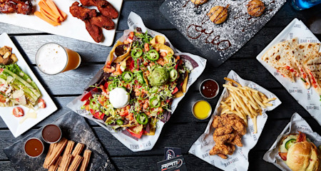 A top view of various dishes on a dark table, including a plate of nachos topped with jalapeños, guacamole, and sour cream, a quesadilla, seasoned french fries, chicken wings, and a glass of beer. Smaller plates hold desserts, fried items, and dipping sauces.