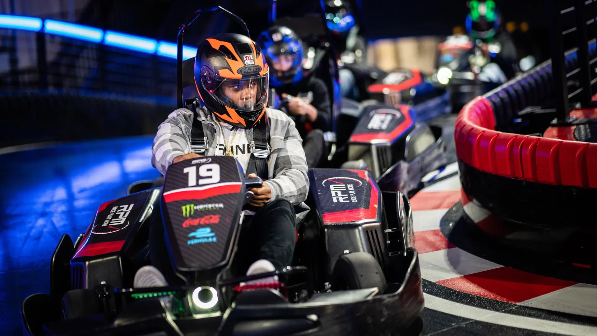 People wearing helmets and driving go-karts are racing on an indoor track. The lead driver, in kart number 19, is navigating a turn. The track is brightly lit with neon lights, and the surrounding is dark with onlookers in the background.