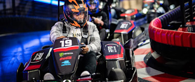 People wearing helmets and driving go-karts are racing on an indoor track. The lead driver, in kart number 19, is navigating a turn. The track is brightly lit with neon lights, and the surrounding is dark with onlookers in the background.