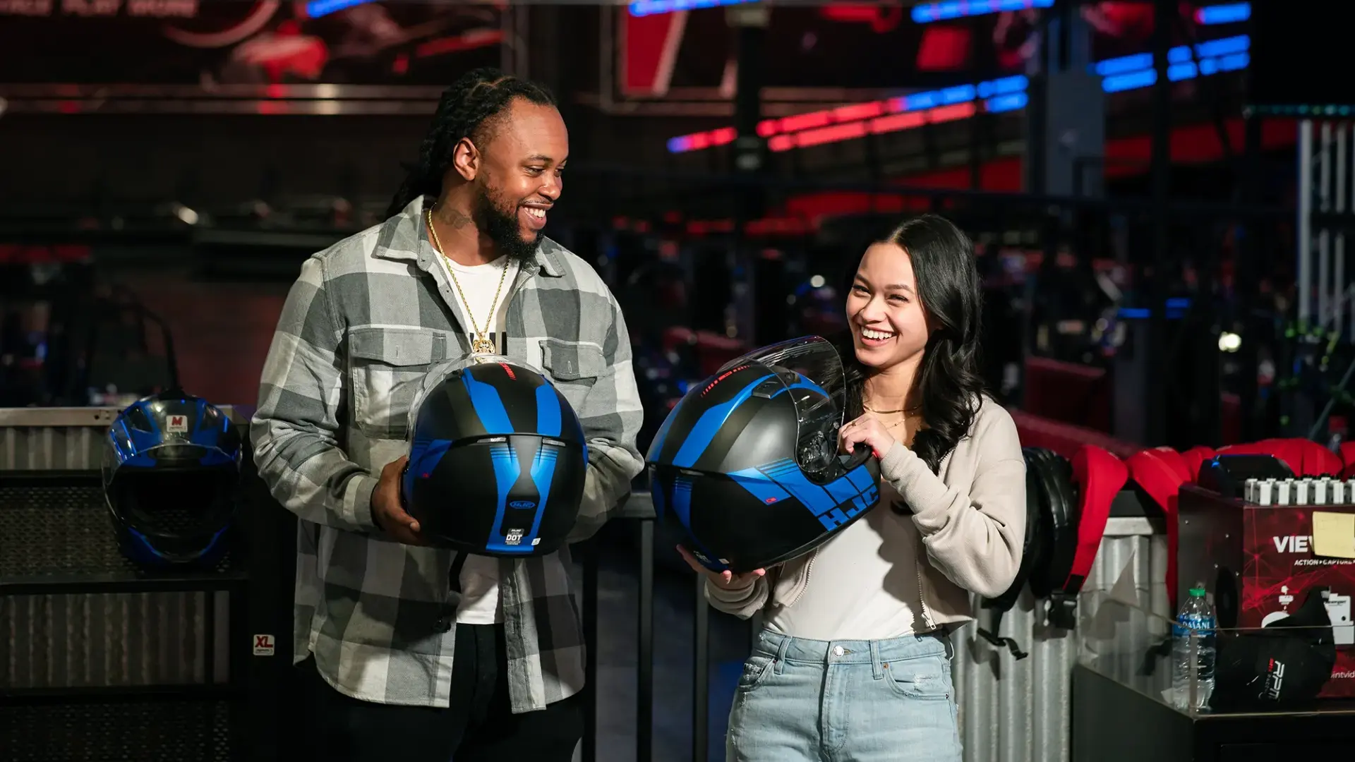 A man and a woman smiling while holding blue and black helmets. They are indoors in a go-karting venue called RPM with other helmets and equipment visible in the background. The man is wearing a checked shirt and the woman is in a gray sweater.