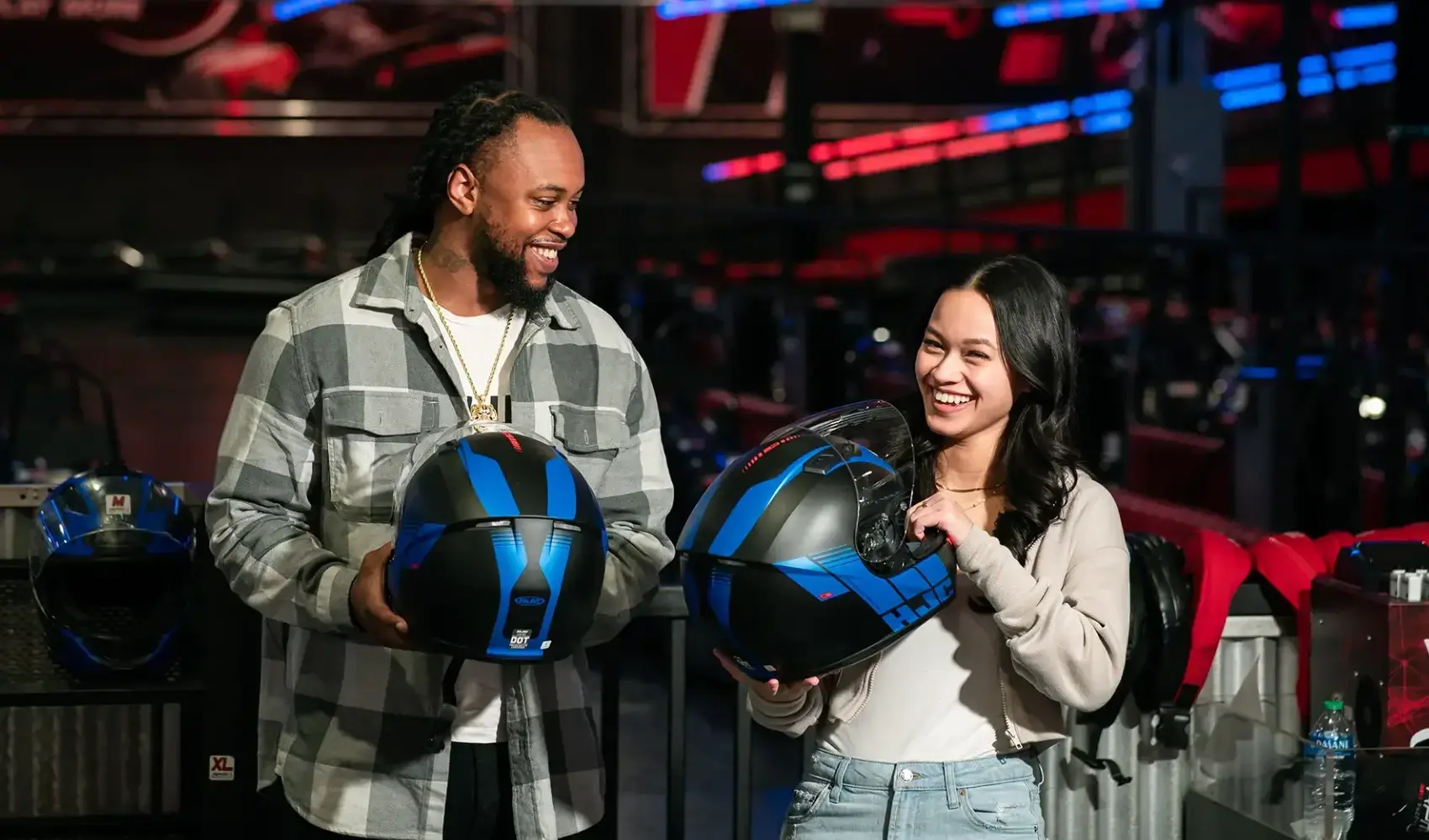 A man and a woman smiling while holding blue and black helmets. They are indoors in a go-karting venue called RPM with other helmets and equipment visible in the background. The man is wearing a checked shirt and the woman is in a gray sweater.