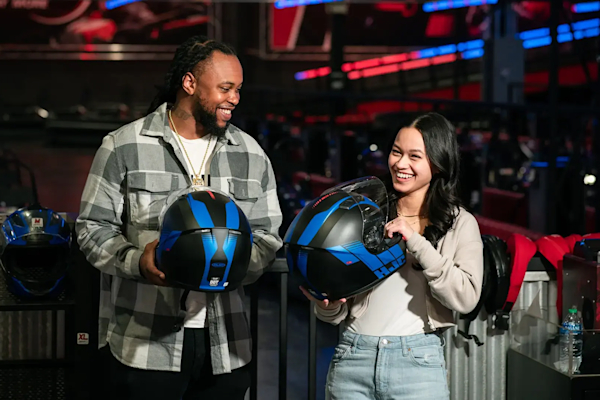 A man and a woman smiling while holding blue and black helmets. They are indoors in a go-karting venue called RPM with other helmets and equipment visible in the background. The man is wearing a checked shirt and the woman is in a gray sweater.