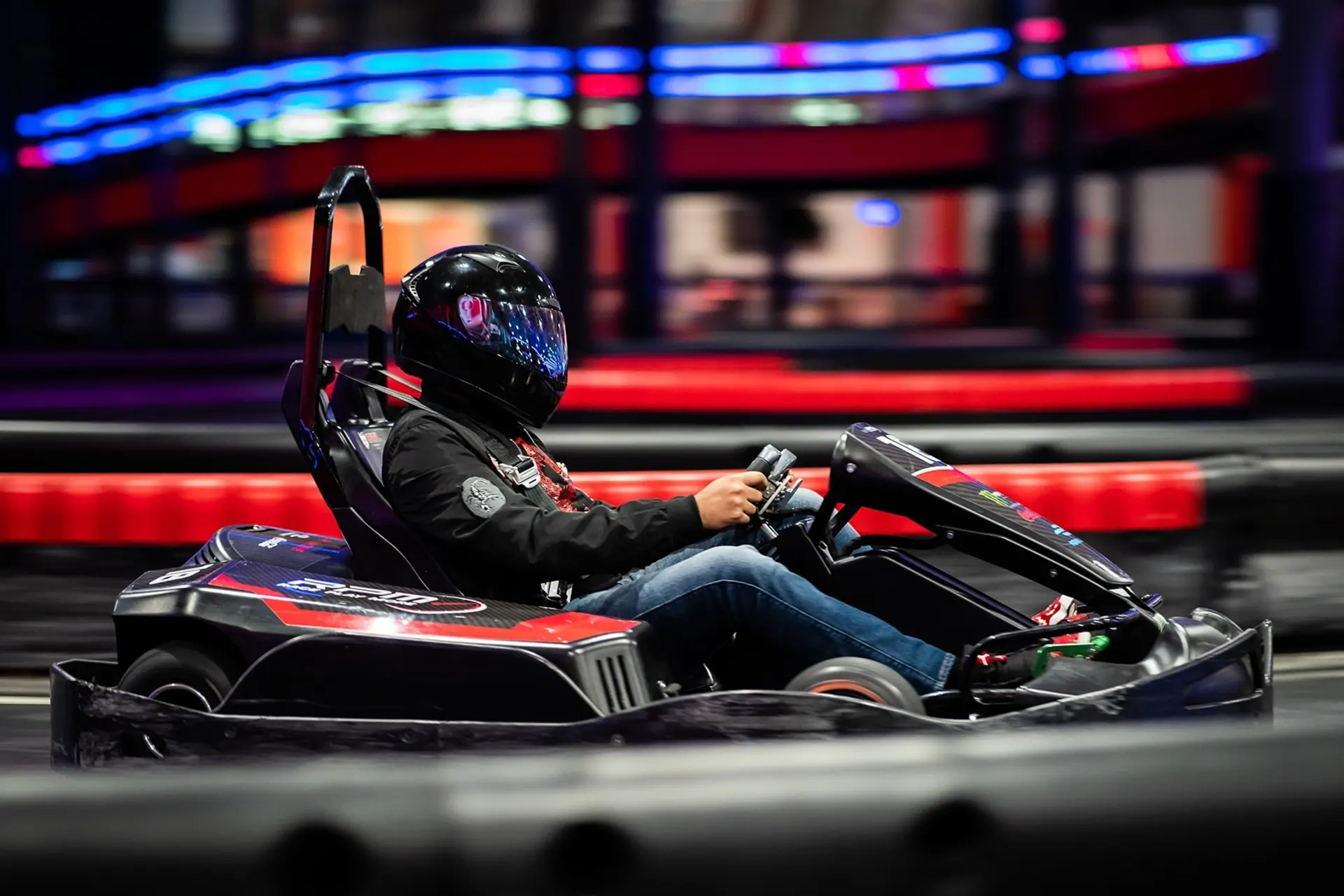 A person wearing a black helmet and a black jacket is driving a go-kart on an indoor track. The background features blurred lights and structures, emphasizing the speed and motion of the scene.