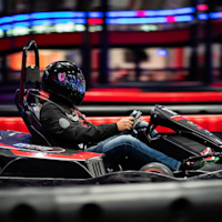 A person wearing a black helmet and a black jacket is driving a go-kart on an indoor track. The background features blurred lights and structures, emphasizing the speed and motion of the scene.