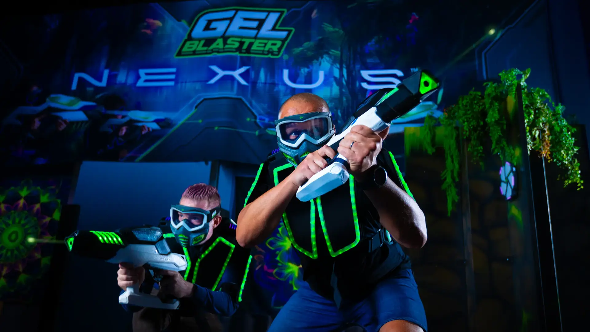 Two people wearing futuristic, glowing vests and protective eyewear, holding gel blasters in a dark, neon-lit arena with a "Gel Blaster Nexus" sign in the background. They appear to be engaged in a lively game or competition.