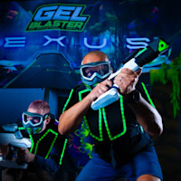 Two people wearing futuristic, glowing vests and protective eyewear, holding gel blasters in a dark, neon-lit arena with a "Gel Blaster Nexus" sign in the background. They appear to be engaged in a lively game or competition.