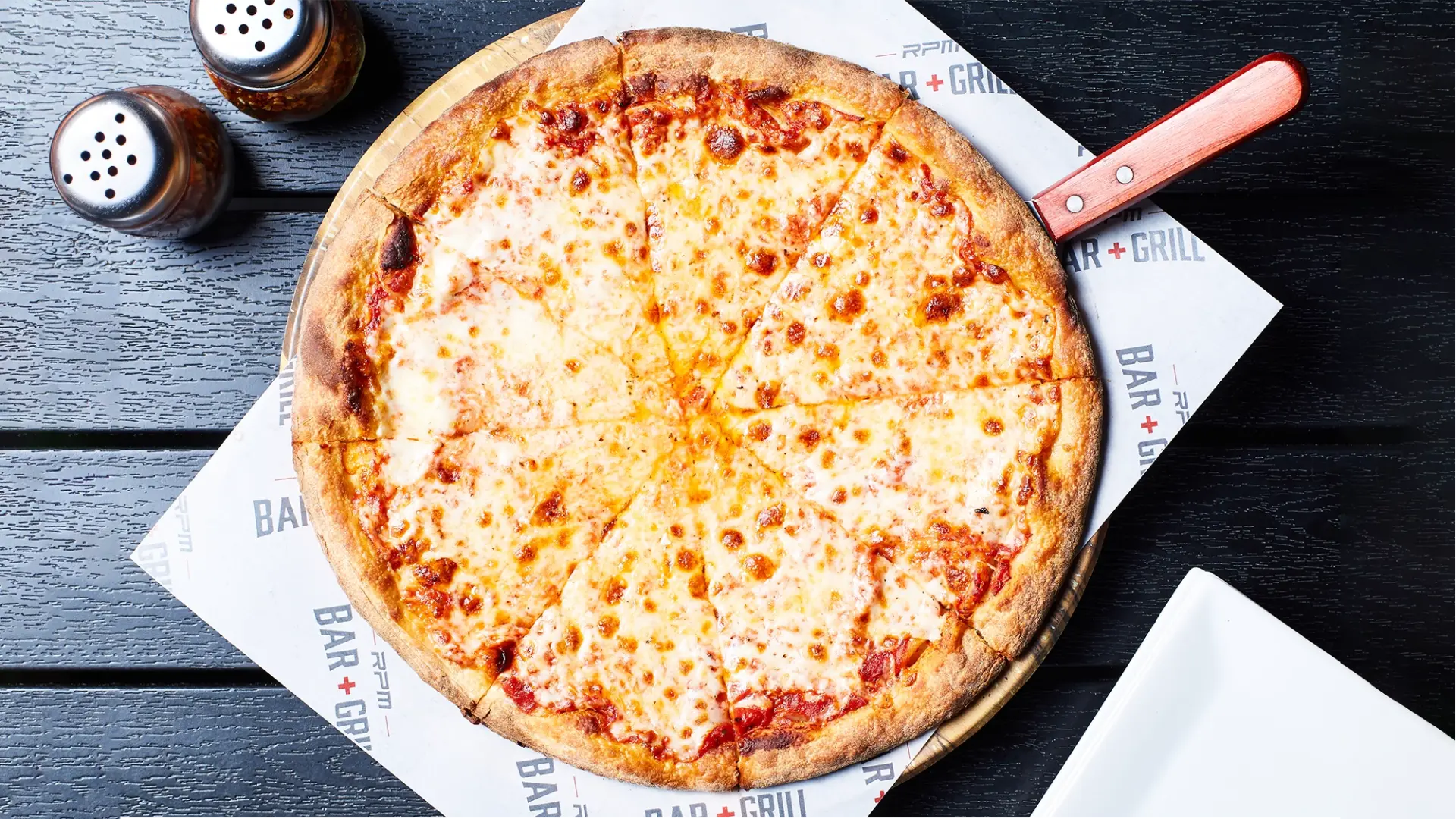 A freshly baked cheese pizza with a golden crust, cut into eight slices, sits on a wooden board. The pizza is placed on branded wax paper that reads " RPM BAR + GRILL". Salt and pepper shakers are visible in the background, along with part of a white plate.