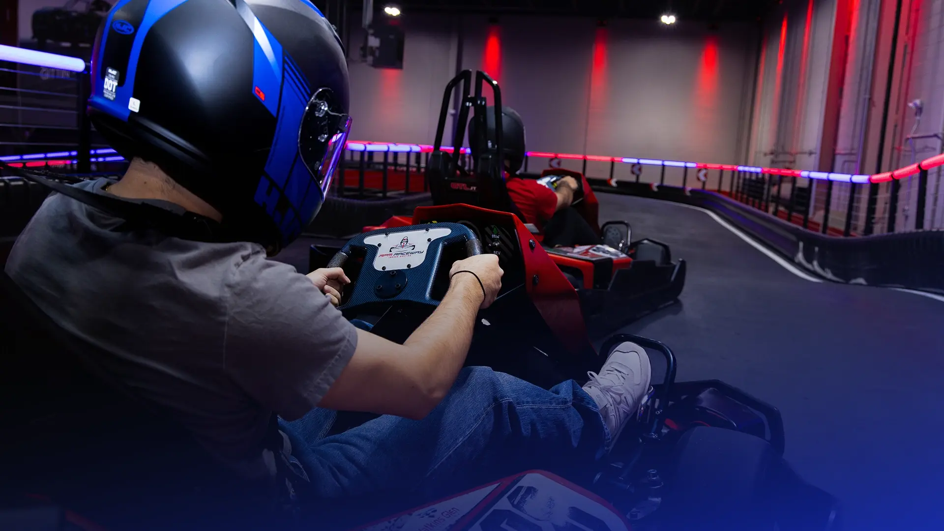 Two people wearing helmets race in go-karts on an indoor track illuminated by red and blue lights. One person is close to the camera, steering the kart while leaning forward. The other person, slightly ahead, is driving another go-kart around a curve.