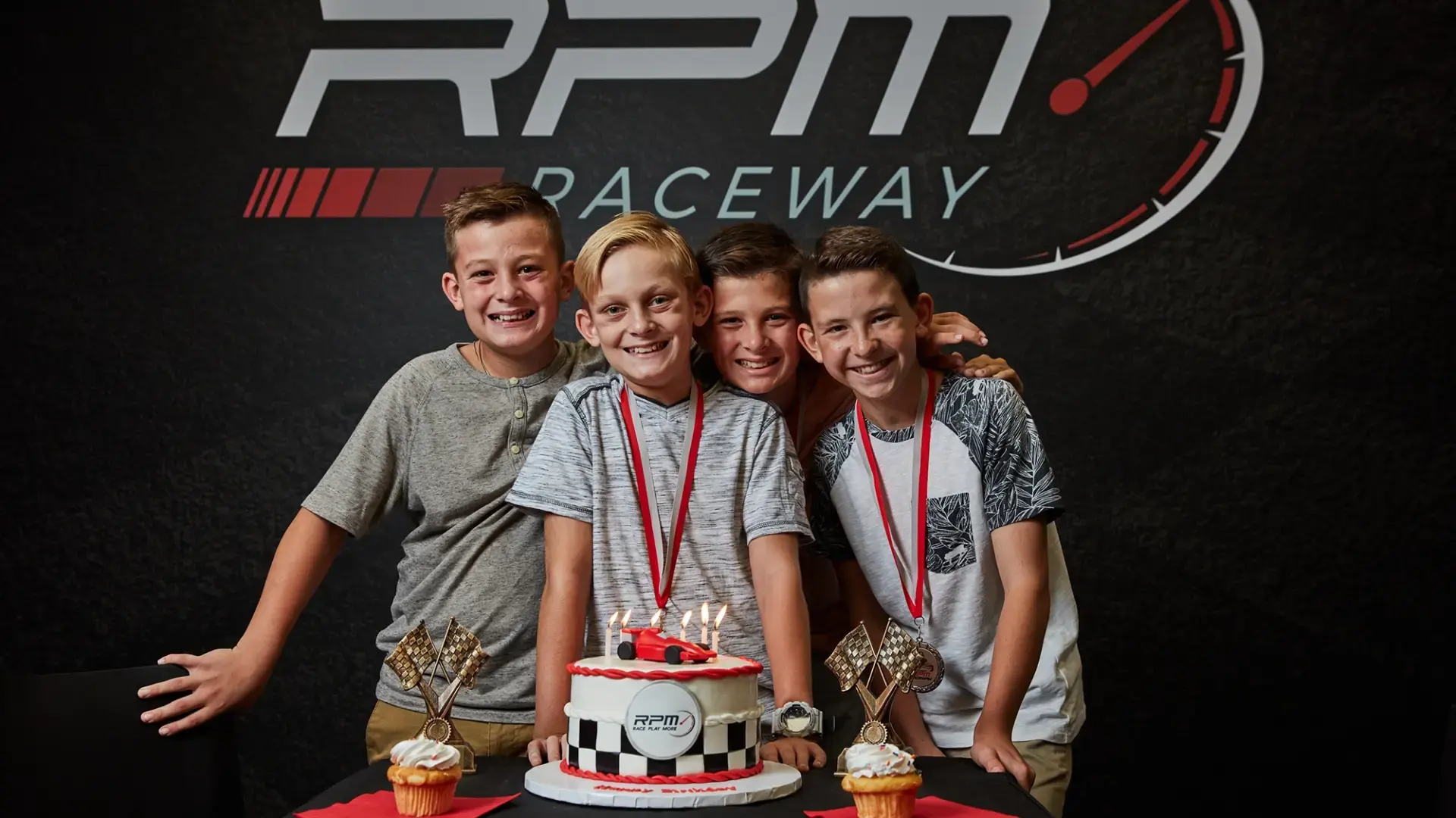 Four boys with medals around their necks smile and pose in front of a racing-themed backdrop that says "RPM Raceway." They are gathered around a table with a decorated cake and two cupcakes, each adorned with a small trophy.