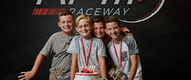 Four boys with medals around their necks smile and pose in front of a racing-themed backdrop that says "RPM Raceway." They are gathered around a table with a decorated cake and two cupcakes, each adorned with a small trophy.