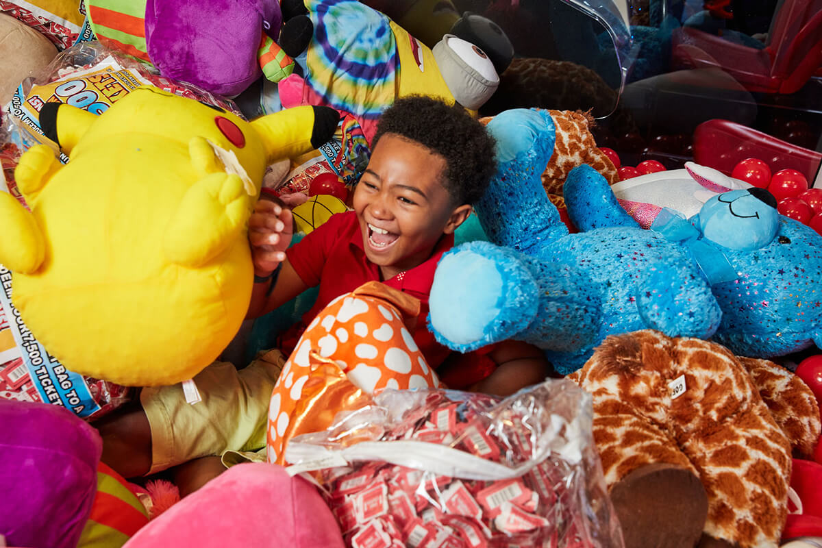 A young child is laughing gleefully while sitting among a large pile of colorful stuffed animals inside a claw machine. The child holds a Pikachu plush and is surrounded by various other soft toys, including a blue bear, a giraffe, and tie-dye patterns.