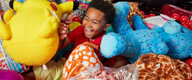 A young child is laughing gleefully while sitting among a large pile of colorful stuffed animals inside a claw machine. The child holds a Pikachu plush and is surrounded by various other soft toys, including a blue bear, a giraffe, and tie-dye patterns.