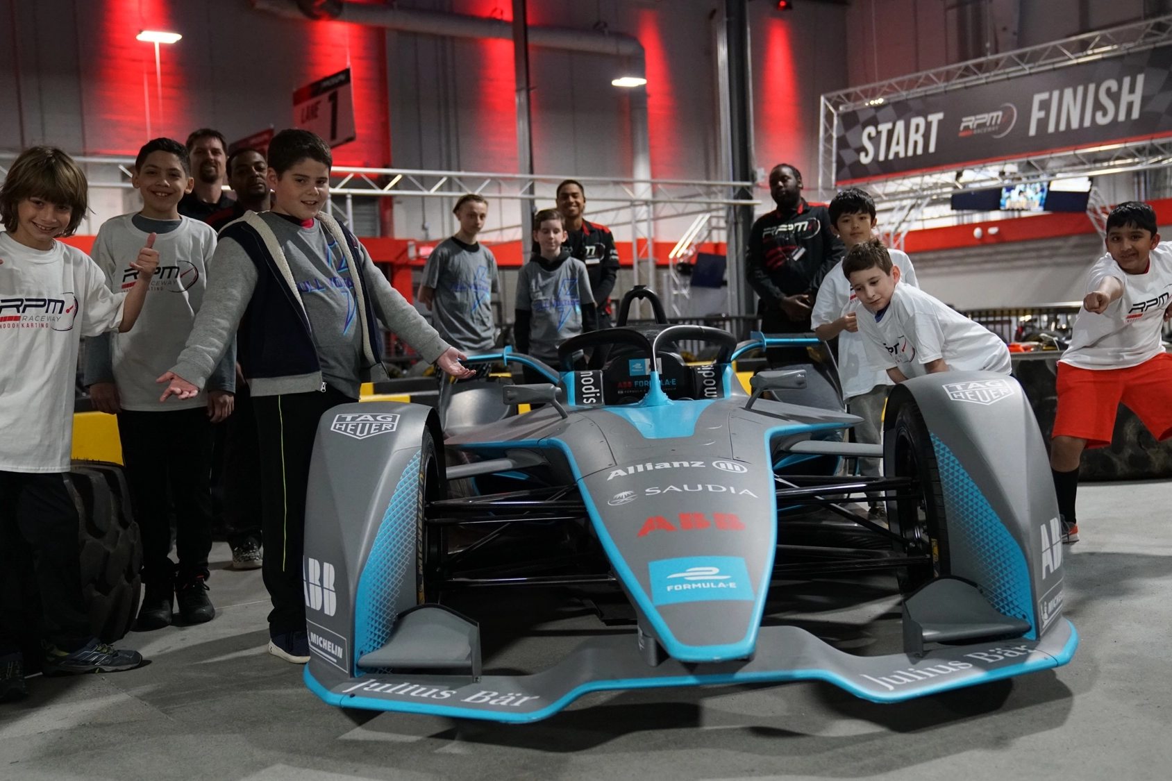 A group of people, including adults and children, pose with smiles and excited expressions around a Formula E race car in an indoor venue at racing camp. The venue has vibrant red lighting, and there is a "START" and "FINISH" banner in the background.