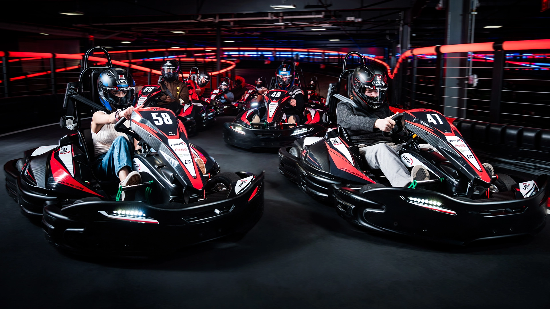 Two people racing go-karts indoors at RPM Raceway. The foreground driver, wearing a green and black helmet, is steering a kart labeled with the number 3. The background driver, in a black and red helmet, follows closely in a kart labeled 18. The track is illuminated with colorful lights.
