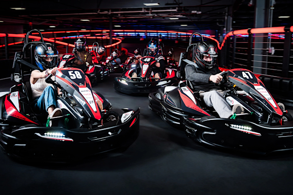 Two people racing go-karts indoors at RPM Raceway. The foreground driver, wearing a green and black helmet, is steering a kart labeled with the number 3. The background driver, in a black and red helmet, follows closely in a kart labeled 18. The track is illuminated with colorful lights.