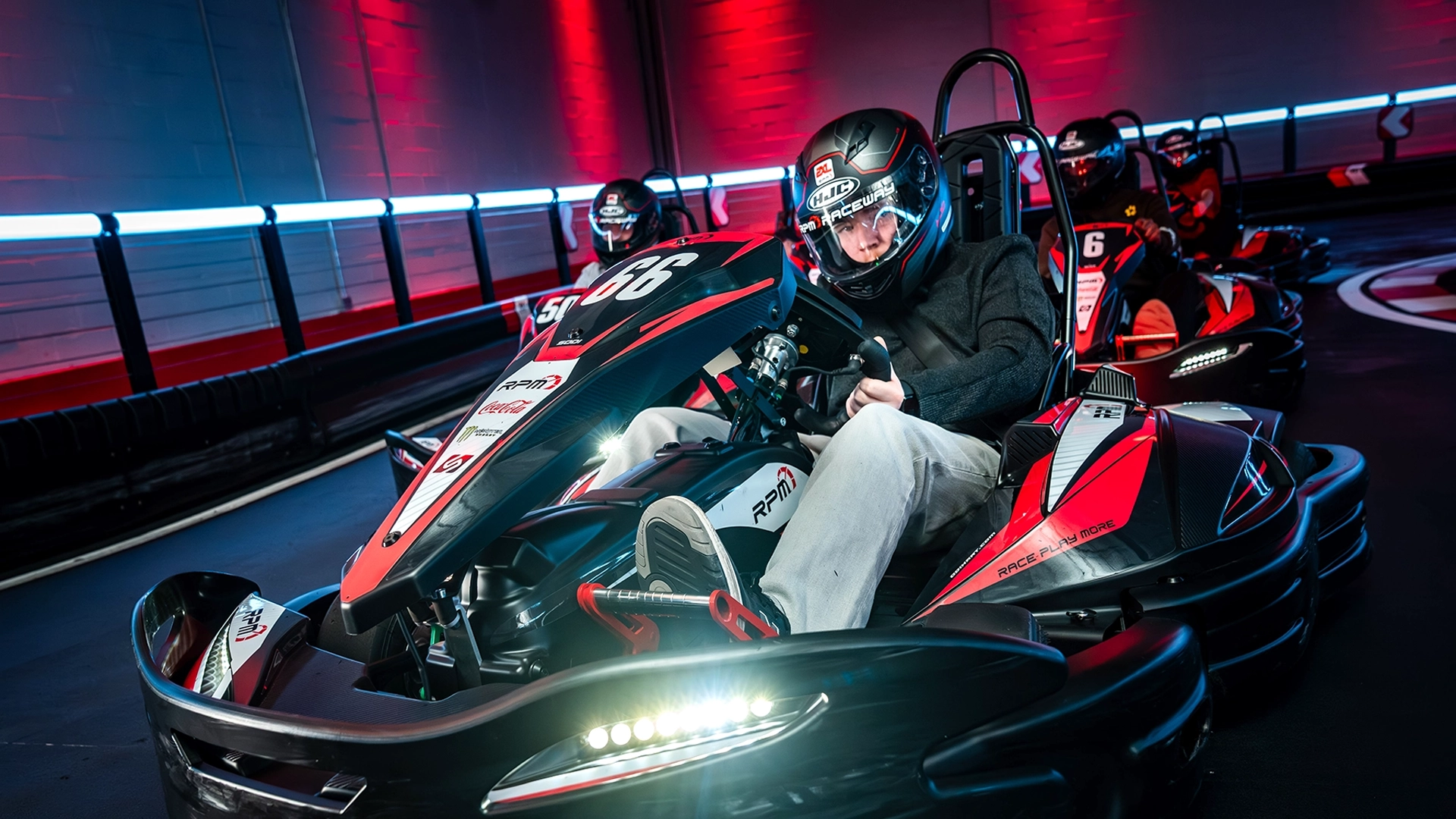 Two people racing go-karts indoors at RPM Raceway. The foreground driver, wearing a green and black helmet, is steering a kart labeled with the number 3. The background driver, in a black and red helmet, follows closely in a kart labeled 18. The track is illuminated with colorful lights.