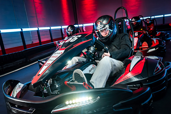 Two people racing go-karts indoors at RPM Raceway. The foreground driver, wearing a green and black helmet, is steering a kart labeled with the number 3. The background driver, in a black and red helmet, follows closely in a kart labeled 18. The track is illuminated with colorful lights.
