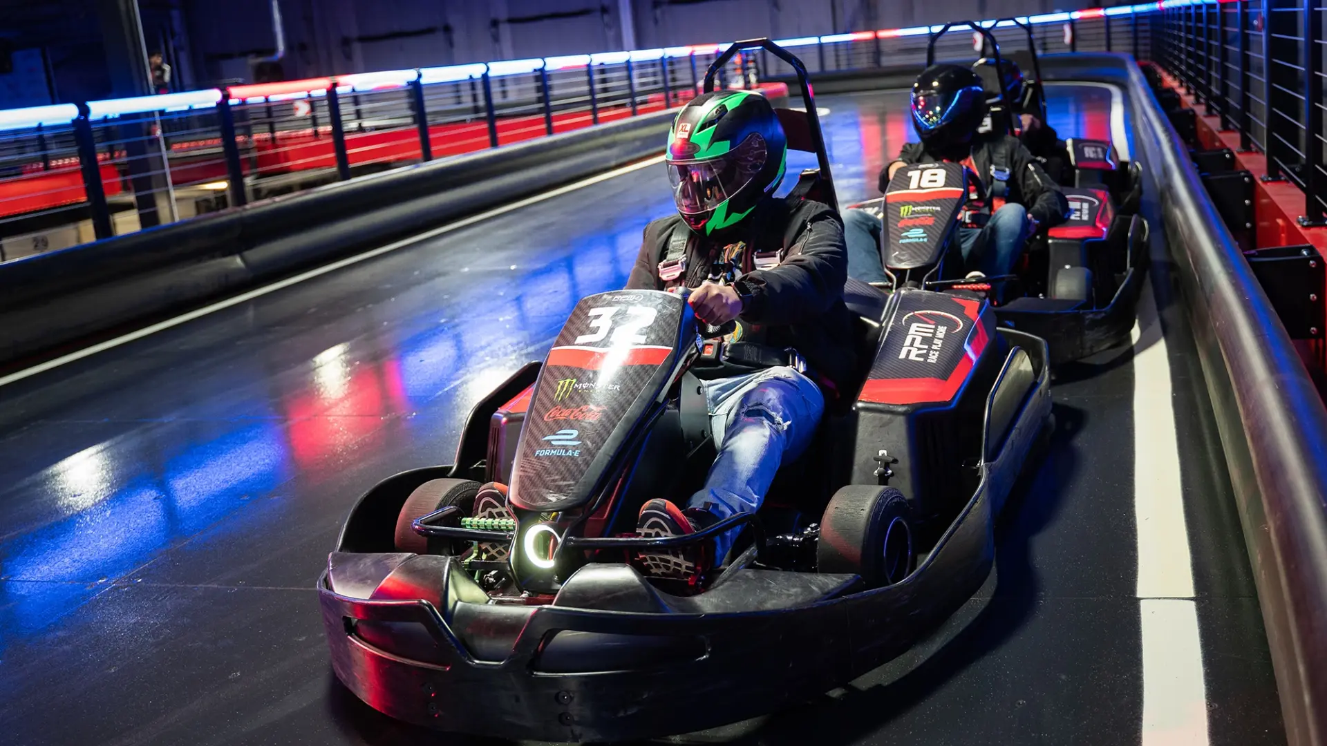 Two people racing go-karts indoors at RPM Raceway. The foreground driver, wearing a green and black helmet, is steering a kart labeled with the number 3. The background driver, in a black and red helmet, follows closely in a kart labeled 18. The track is illuminated with colorful lights.