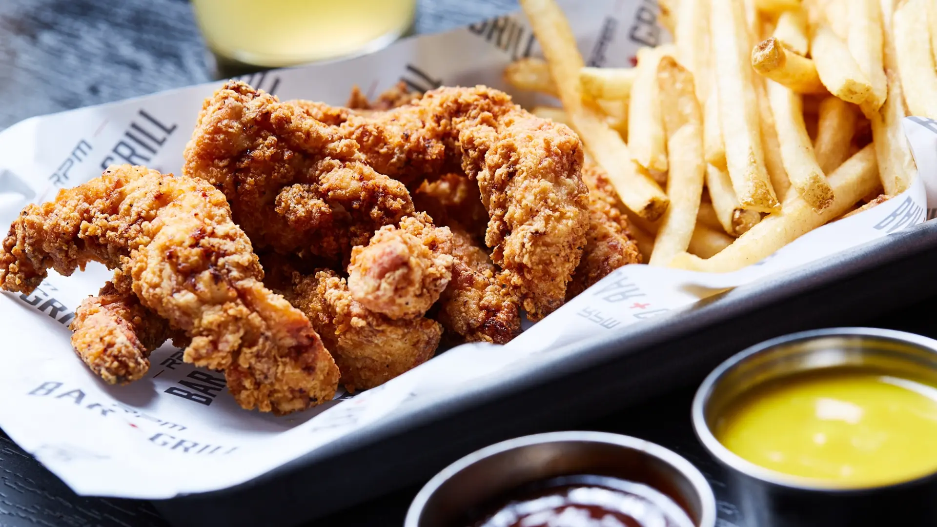 A close-up of a meal at RPM Bar + Grill featuring crispy fried chicken tenders and golden french fries served on a rectangular tray lined with branded paper. In the foreground, two dipping sauces, one yellow and one brown, are placed in small metal ramekins. A drink is blurred in the background.