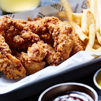 A close-up of a meal at RPM Bar + Grill featuring crispy fried chicken tenders and golden french fries served on a rectangular tray lined with branded paper. In the foreground, two dipping sauces, one yellow and one brown, are placed in small metal ramekins. A drink is blurred in the background.