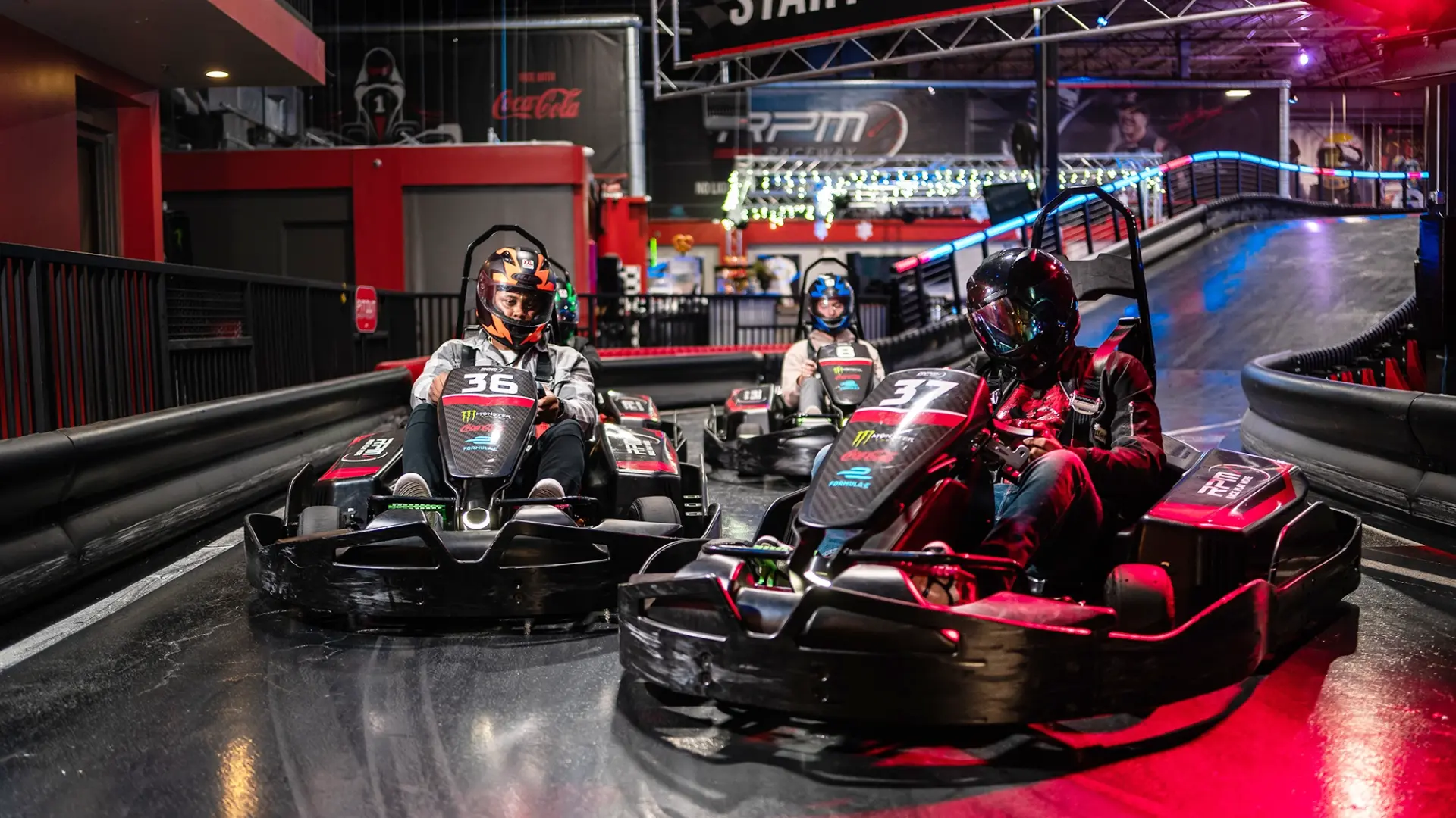 Three individuals are racing go-karts indoors. The racers are wearing helmets and each kart is marked with a number (36 on the left, 31 on the right). The track has barriers to guide the route, and the background shows a colorful, well-lit arcade area.