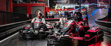 Three individuals are racing go-karts indoors. The racers are wearing helmets and each kart is marked with a number (36 on the left, 31 on the right). The track has barriers to guide the route, and the background shows a colorful, well-lit arcade area.
