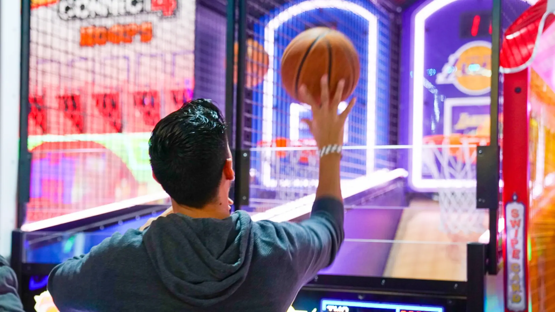 A person stands at an arcade basketball game, poised to shoot a basketball towards the hoop. The brightly lit game has multiple hoops and a digital score display. The background is colorful with various arcade decorations and lights.