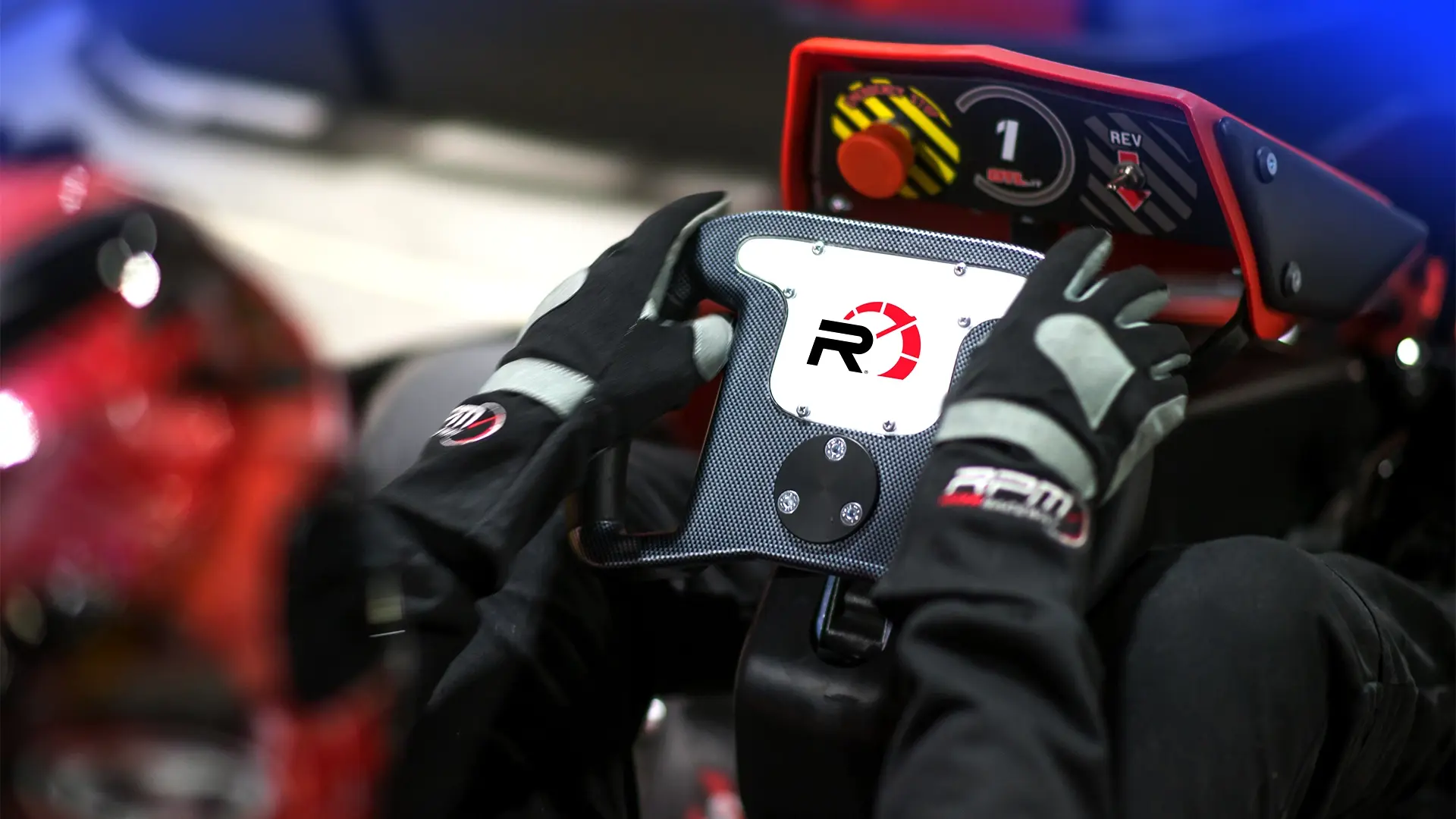 Close-up of a gloved person holding a racing steering wheel, with a digital screen and controls in the background. The steering wheel features the logo "R1". The atmosphere suggests a high-tech racing environment, possibly a simulation or an actual race car.