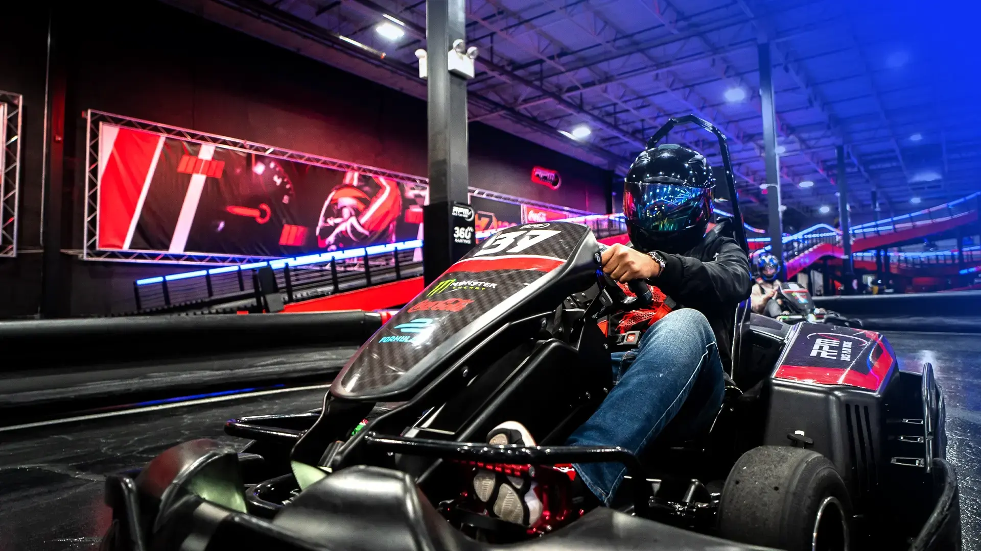 A person wearing a blue helmet and black clothing drives a black go-kart on an indoor track with vibrant red and blue lighting. The person is holding the steering wheel, focusing on the track ahead. Colorful wall designs and racing decorations are visible in the background.