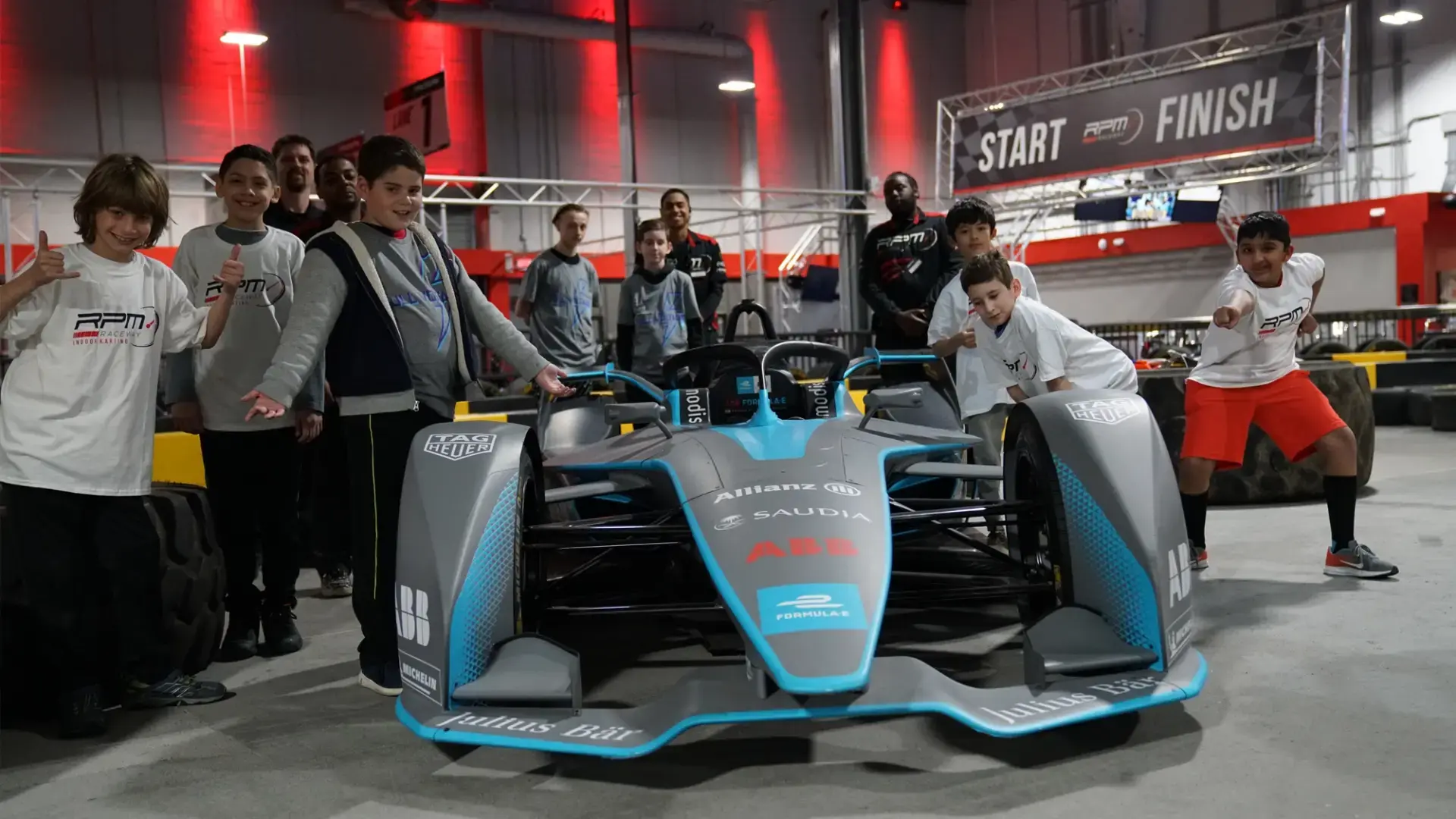 A group of people, including adults and children, pose with smiles and excited expressions around a Formula E race car in an indoor venue at racing camp. The venue has vibrant red lighting, and there is a "START" and "FINISH" banner in the background.