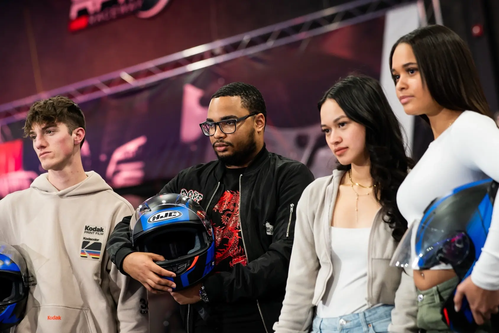 Four people stand together indoors, each holding or wearing a motorcycle helmet. They appear to be at a racing event or exhibition, with a blurred background featuring a stage or display area.