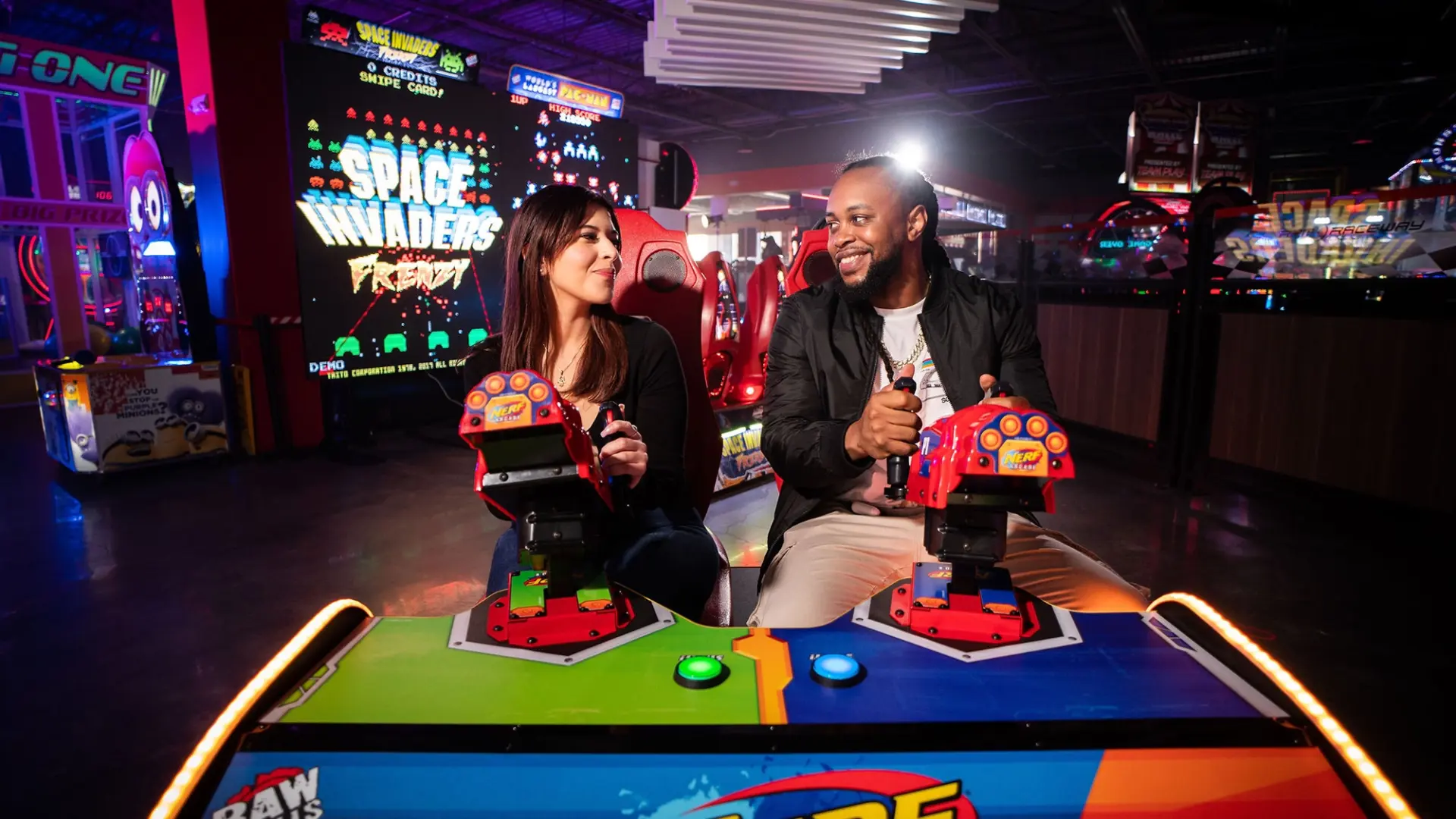 Two people are sitting side by side at an arcade game, each holding a gun controller and smiling at each other. The background shows brightly lit arcade machines and a sign that reads "Space Invaders." The atmosphere is lively and colorful