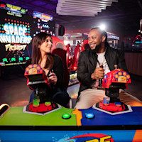 Two people are sitting side by side at an arcade game, each holding a gun controller and smiling at each other. The background shows brightly lit arcade machines and a sign that reads "Space Invaders." The atmosphere is lively and colorful