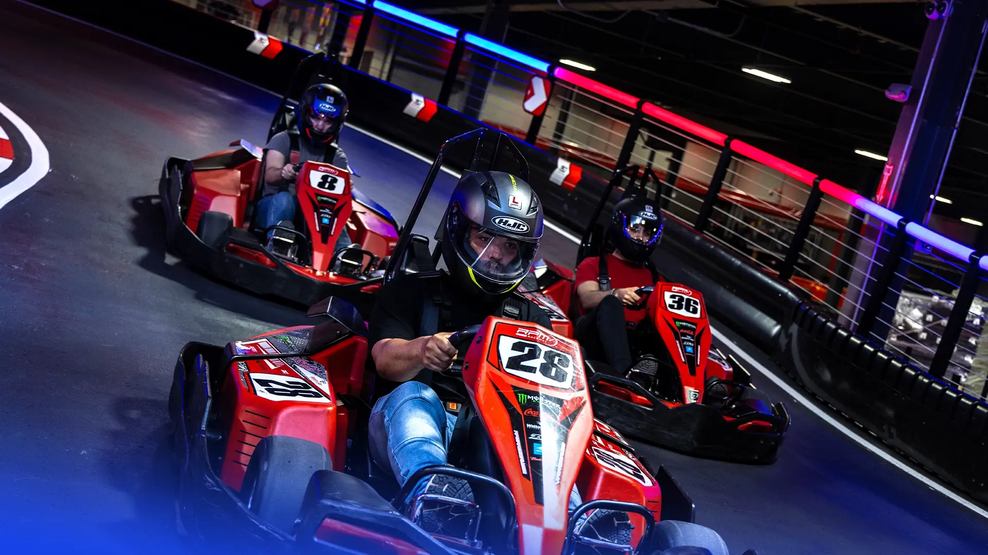 Three individuals are racing go-karts on an indoor track. They are wearing helmets and driving red go-karts. The foremost driver is in kart number 28, followed by kart number 8 and kart number 36. Colorful lights illuminate the track's surroundings.