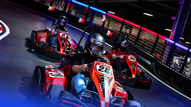 Three individuals are racing go-karts on an indoor track. They are wearing helmets and driving red go-karts. The foremost driver is in kart number 28, followed by kart number 8 and kart number 36. Colorful lights illuminate the track's surroundings.