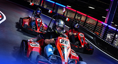 Three individuals are racing go-karts on an indoor track. They are wearing helmets and driving red go-karts. The foremost driver is in kart number 28, followed by kart number 8 and kart number 36. Colorful lights illuminate the track's surroundings.