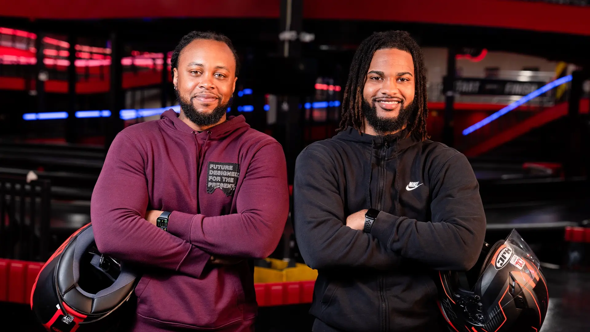 Two men are standing side by side in a go-kart facility called rpm raceway, each holding a helmet under one arm. Both are smiling and have their arms crossed. One wears a maroon hoodie with text on it, the other wears a black Nike hoodie. The setting appears colorful and dynamic.