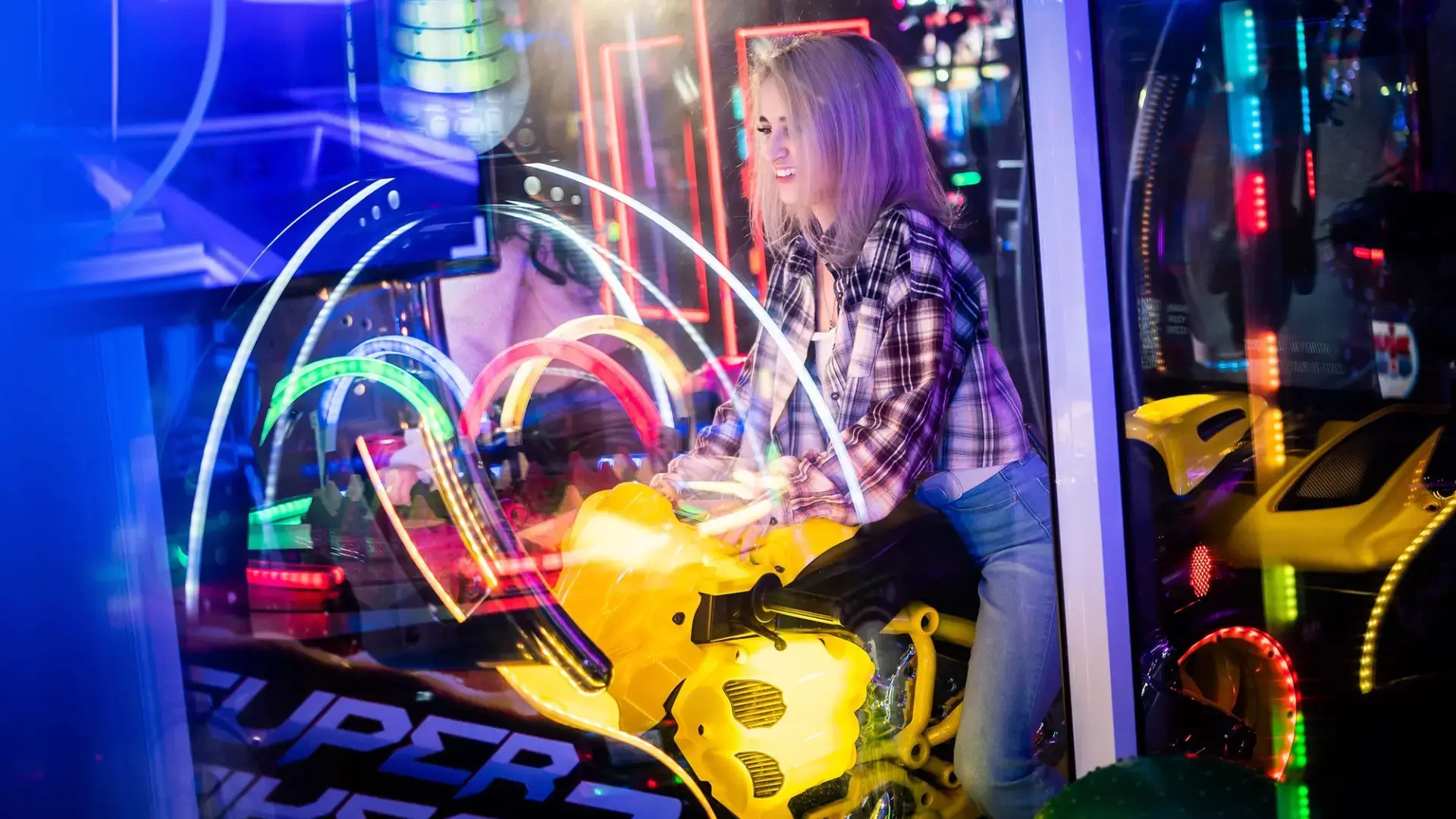 A person rides a yellow motorbike arcade game in a vibrant, neon-lit gaming center. The background is filled with colorful lights and reflections, enhancing the lively atmosphere. They appear to be enjoying the immersive gaming experience.