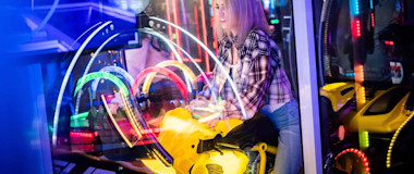 A person rides a yellow motorbike arcade game in a vibrant, neon-lit gaming center. The background is filled with colorful lights and reflections, enhancing the lively atmosphere. They appear to be enjoying the immersive gaming experience.