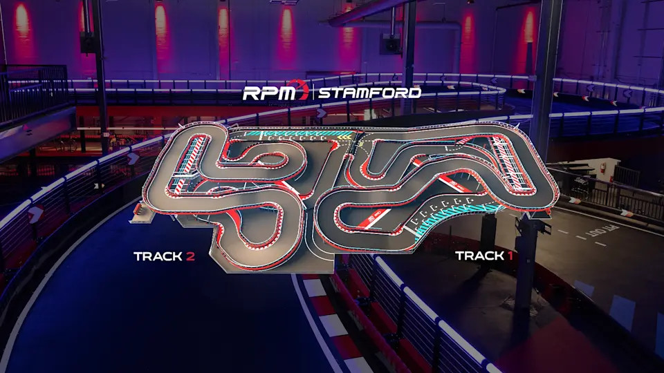 A complex indoor go-kart track with multiple winding turns, lit under blue and red lights. The track layout features several straightaways and sharp corners, designed for a challenging racing experience. The background includes part of the racing facility with additional lighting.