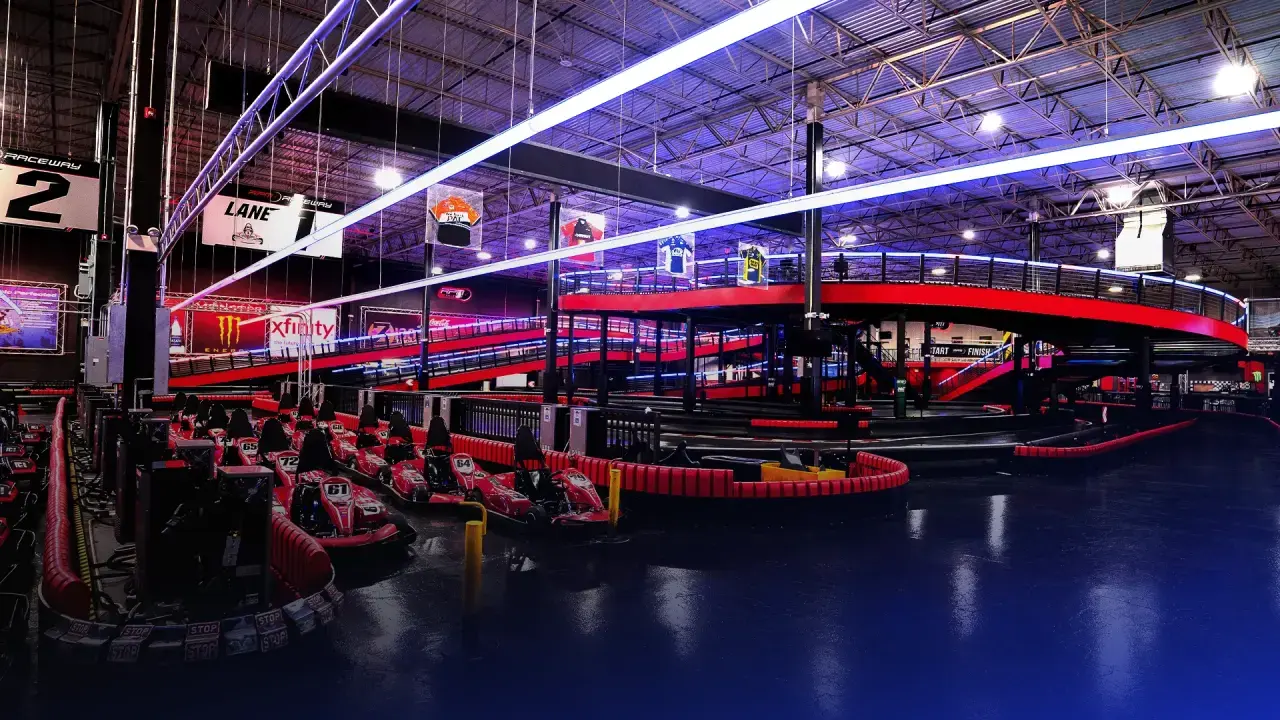 Indoor go-kart racing track with multiple red karts lined up under bright neon and LED lights. The track features winding curves, safety barriers, and an elevated section with a ramp. The atmosphere is energetic and futuristic.