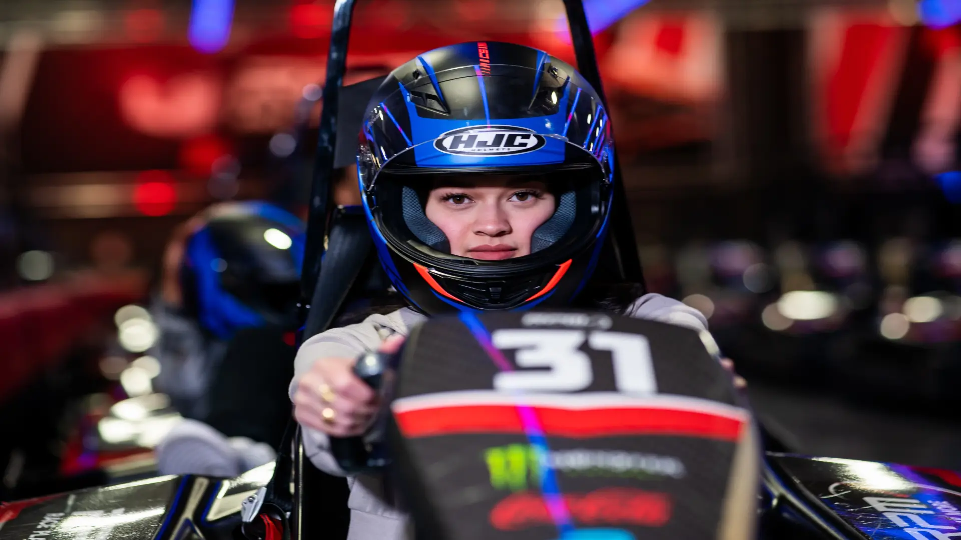 A person wearing a black racing helmet with a blue visor, and a gray racing suit sits in a go-kart, gripping the steering wheel. The number 31 is prominently displayed on the front of the kart, along with various decals. The background is blurred with red and neon lights.