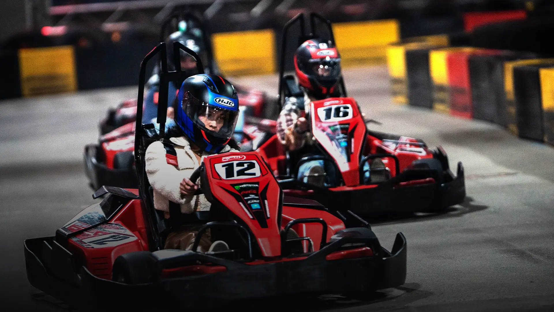 Children in protective gear race go-karts on an indoor track. The front racer, wearing helmet number 12, speeds ahead of the others. The track is surrounded by colorful barriers. The scene is energetic and competitive.