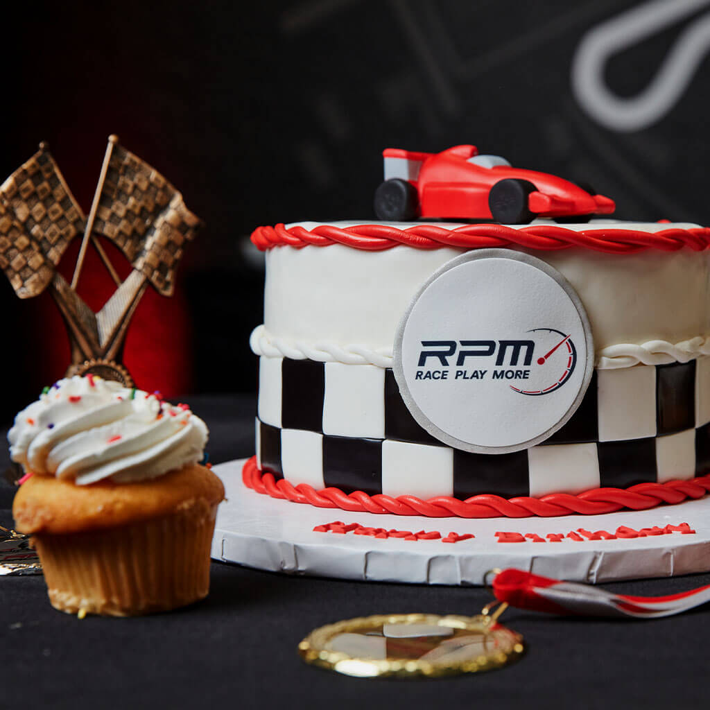 A race car-themed cake with a red and black checkered design, featuring a small red race car on top. Next to the cake is a cupcake with white frosting, a trophy with crossed checkered flags, and a medal. The cake has "RPM" and "Race Play More" printed on it.