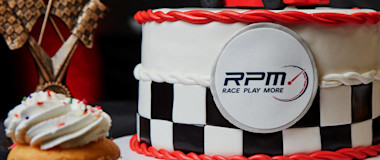 A race car-themed cake with a red and black checkered design, featuring a small red race car on top. Next to the cake is a cupcake with white frosting, a trophy with crossed checkered flags, and a medal. The cake has "RPM" and "Race Play More" printed on it.