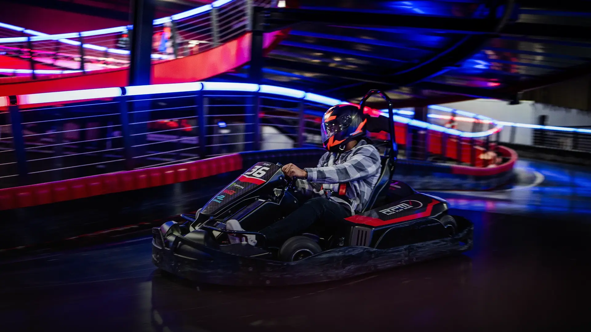 A person wearing a helmet and racing gear is driving a go-kart on an indoor track with a multi-level design. Neon lights create a vibrant atmosphere, highlighting the dynamic motion of the go-kart as it speeds through a curve.