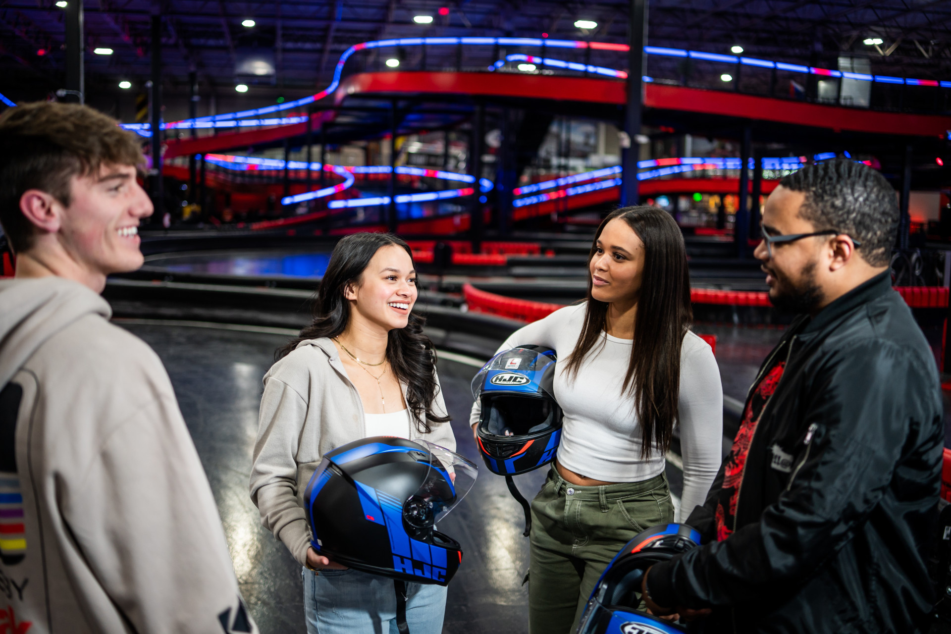 Four people, three women and one man, standing and chatting indoors. They are holding helmets, suggesting they are at a go-karting track. Colorful lights and the racing tracks are visible in the background. All appear to be smiling and engaged in conversation.