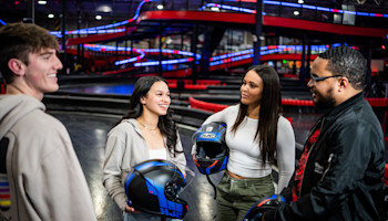 Four people, three women and one man, standing and chatting indoors. They are holding helmets, suggesting they are at a go-karting track. Colorful lights and the racing tracks are visible in the background. All appear to be smiling and engaged in conversation.